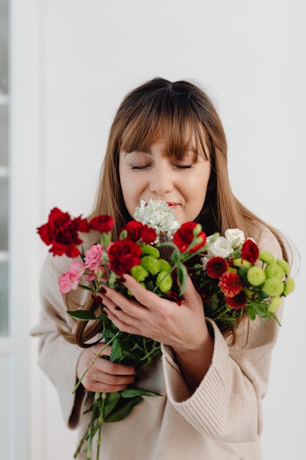 A young woman with light brown hair and bangs, dressed in a beige sweater, is holding a vibrant bouquet of fresh flowers close to her face with both hands, gently pressing the blossoms against her che