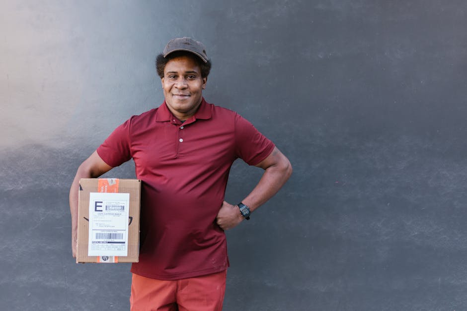 A smiling delivery person with dark skin and Short curly hair, wearing a maroon polo shirt, matching pants, and a dark cap, is standing against a plain dark grey wall. He is holding a small cardboard